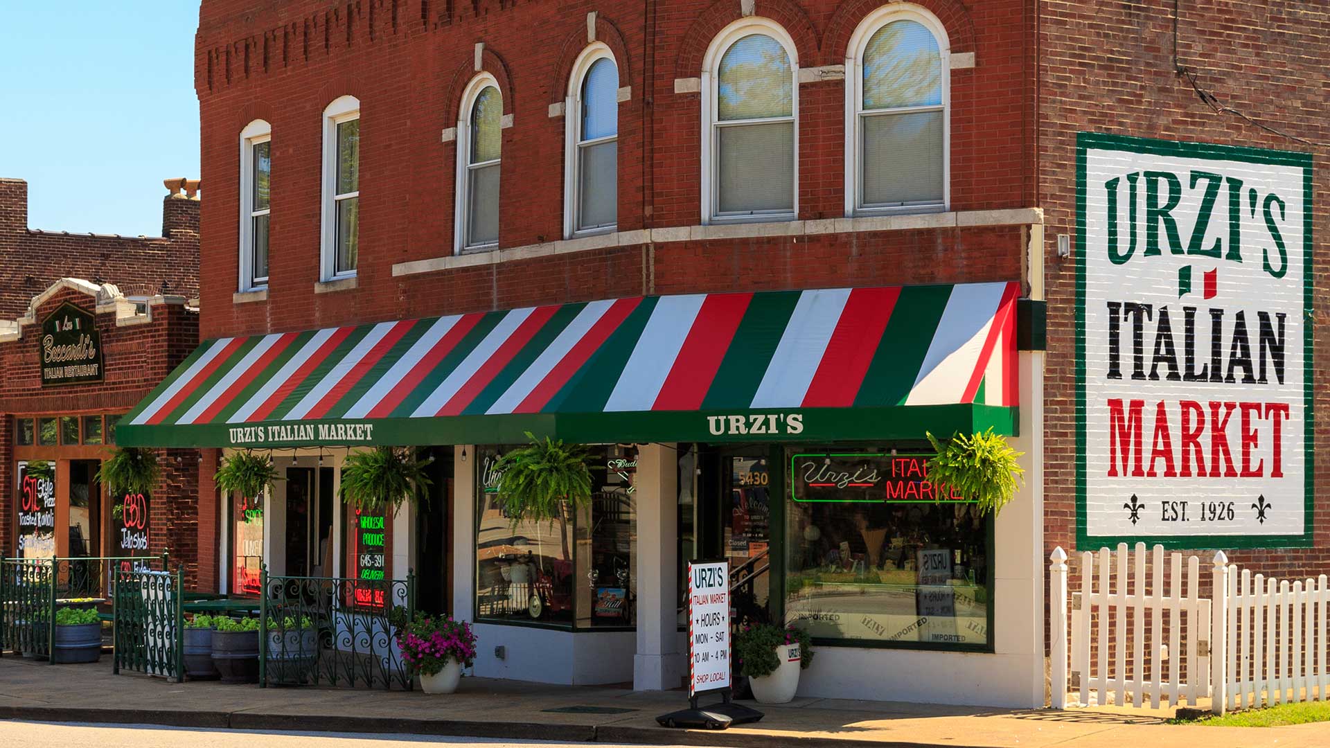 An Italian market in The Hill district of St. Louis. A red, green and white awning hangs out front with several potted plants set along the sidewalk.
