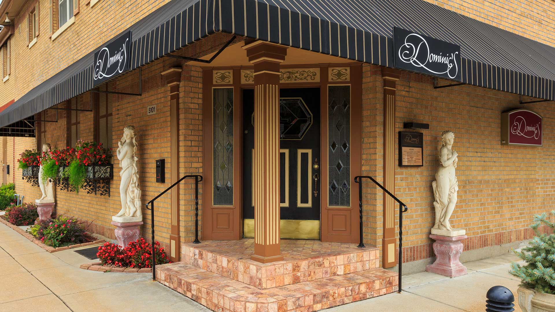 The front door of a classic Italian restaurant. The building is yellow brick with a black awning hanging. There are old-world statues and flowers lining the walls on both sides of the entrance.