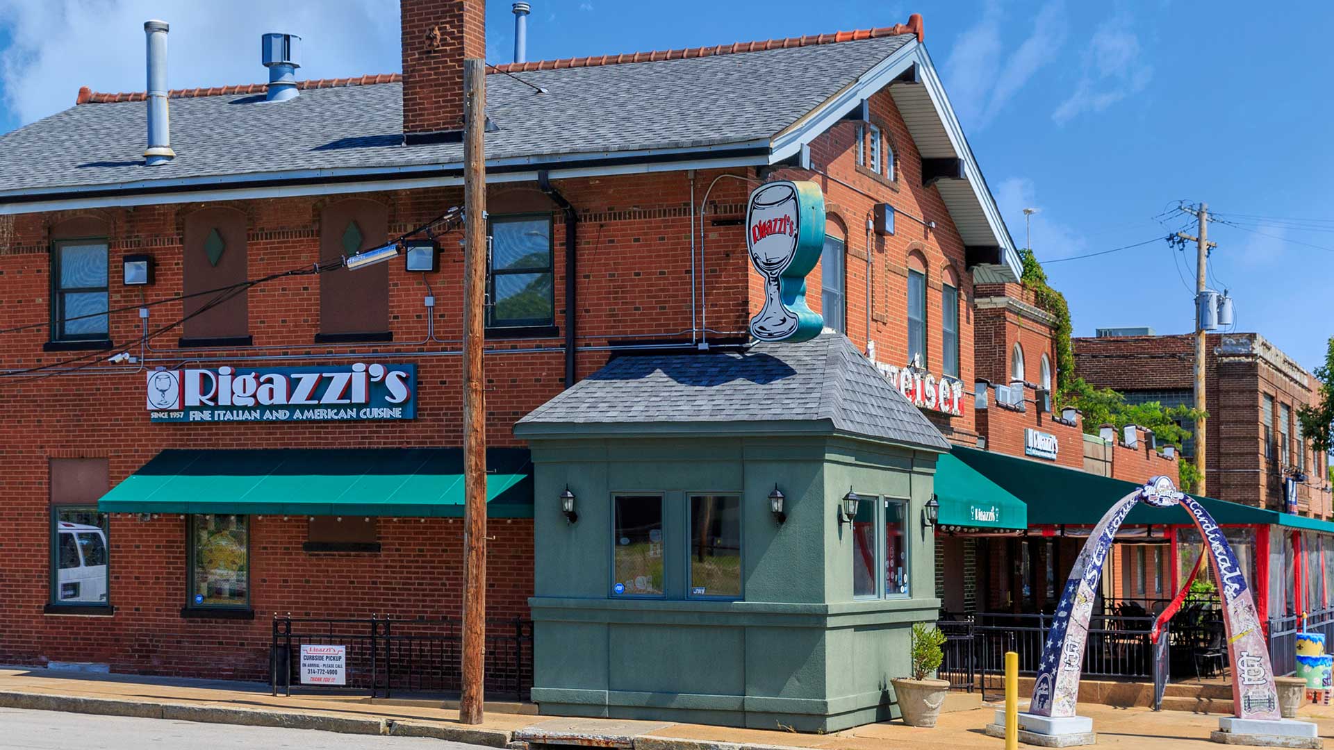 The exterior of a restaurant with the red brick building to the left and an outdoor seating area to the right. A miniature of the St. Louis Gateway Arch is in front.