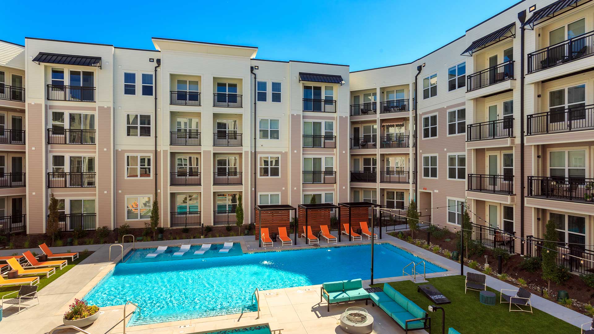 Looking at the amenity courtyard from above. The pool is below, surrounded by lounge chairs. There is outdoor couches and a game area just below to the right. The apartment building surrounds the scene.