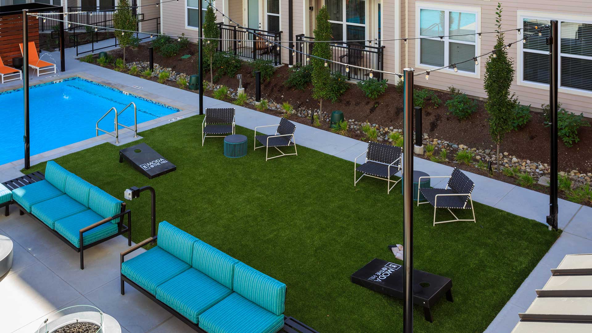 Looking at the outdoor gaming area from above. There is green turf with a cornhole set. There is seating on both sides. The pool is just off to the left.