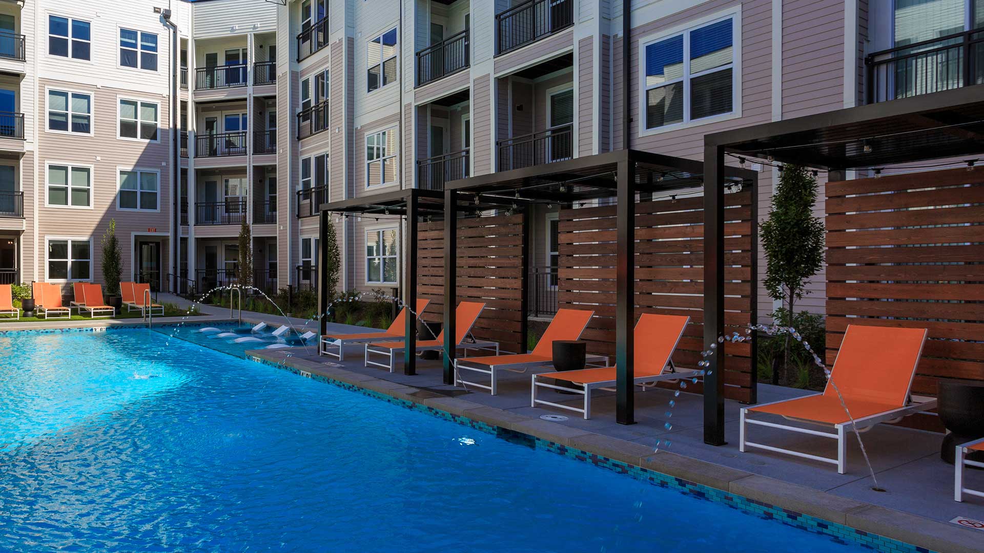 Looking across the pool at the poolside cabanas with orange lounge chairs beneath. The apartment building is in the background.