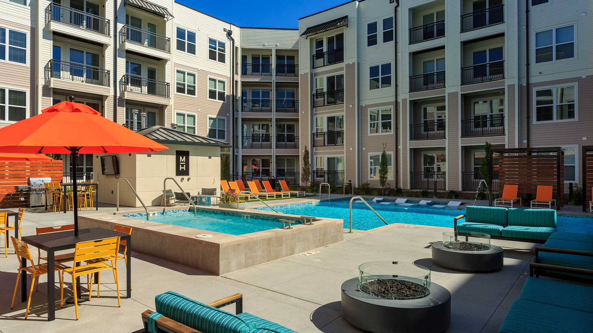 Standing in the courtyard looking across a sitting are with fire pits. The hot and pool are ahead with a grilling and seating area off to the left. The apartment building is seen in the background.